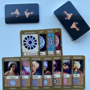 May include: A set of 10 tarot cards with gold frames and intricate designs. The cards feature images of people and geometric patterns. The cards are laid out in a row on a white surface.
