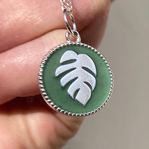 May include: A silver necklace with a green and white enamel pendant featuring a monstera leaf design.