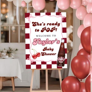 May include: A baby shower sign on an easel with the text "She's ready to POP!" and "Welcome to Taylor's Baby Shower." The sign features a Dr. Pepper bottle and cherries. Balloons in shades of pink and red surround the sign.