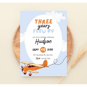 以下が含まれることがあります： オレンジと青で「THREE years FLEW BY」の文字が入った水色の誕生日招待状。「Hudson」の名前、日付、時間、住所が記載されており、オレンジ色の飛行機のイラストが特徴です。
