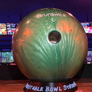 May include: A green bowling ball with the word "Brunswick" in white lettering. The ball has a large finger hole and is resting on a black stand that reads "Irritable Bowl Syndrome" in white.