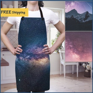May include: A blue and purple galaxy print apron with a black adjustable strap. The apron is worn by a person in a white shirt. The background is a kitchen with white cabinets and a window.