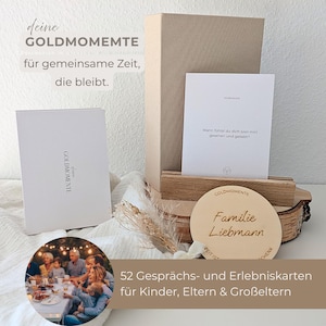 Puede incluir: Un juego de regalo con una caja blanca, una tarjeta y un letrero redondo de madera. El letrero dice "Familie Liebmann". El juego incluye 52 tarjetas de conversación y experiencia para niños, padres y abuelos. El texto "deine GOLDMOMENTE" también es visible.