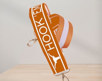 May include: Orange and white striped adjustable strap with the text "HOOK 'EM" and a white silhouette of a longhorn steer. The strap has gold metal hardware.