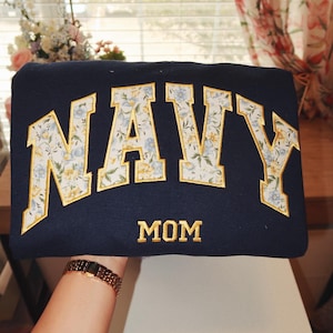 May include: Navy blue sweatshirt with the word "NAVY" in large, floral-patterned letters outlined in gold. The word "MOM" is embroidered in gold below the word "NAVY".