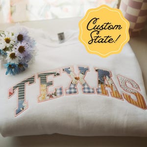 May include: White sweatshirt with a patchwork "TEXAS" design in pink, blue, and yellow. The sweatshirt is folded and has a yellow tag that says "Custom State!"