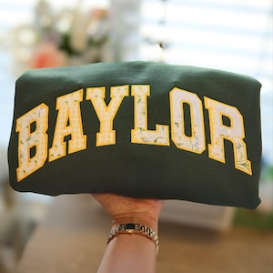 May include: Dark green pillow with the word "BAYLOR" in large, yellow, block letters. The letters have a white outline and are filled with a floral and gingham pattern. The pillow is held up by a person.