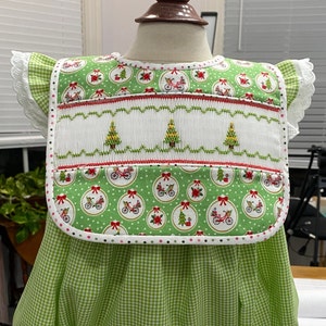 May include: A green and white gingham dress with a white smocked bib featuring a red and green Christmas tree design. The bib has a green, white, and red trim.