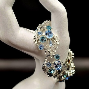 May include: A silver-toned bracelet with blue and teal gemstones is displayed on a white hand-shaped jewelry holder. The bracelet features a floral design with multiple gemstones. The background is black.