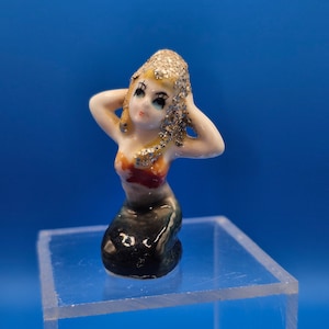 May include: A small ceramic mermaid figurine with blonde hair covered in glitter, arms raised to her head. The mermaid has a red and orange top and a black tail. The figurine is kneeling on a clear acrylic block against a blue background.