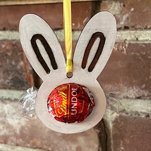 May include: White wooden bunny rabbit cutout with a red Lindor chocolate truffle inside. The bunny is hanging from a yellow ribbon.