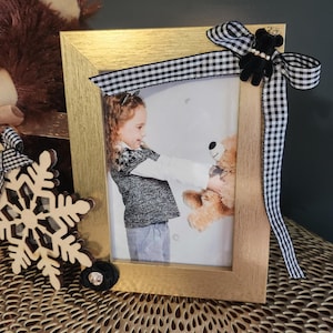 May include: A gold picture frame with a black and white checkered ribbon and a black bow with a small black teddy bear charm. The frame contains a photo of a young girl holding a teddy bear.