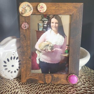 May include: A brown wooden picture frame with a photo of a person holding a white rabbit. The frame is decorated with four buttons, two pink, one beige, and one with a floral design.