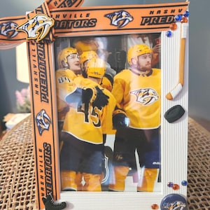 May include: A white picture frame with a hockey theme. The frame is decorated with orange and black ribbon with the words "Nashville Predators" printed on it. The frame also has a hockey stick, a puck, and a hockey skate. The picture inside the frame shows three hockey players wearing yellow and blue jerseys.