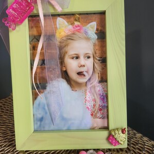 Puede incluir: Un marco de fotos verde con una foto de una niña pequeña con una diadema de unicornio. El marco está decorado con una cinta rosa y un encanto de cupcake rosa con purpurina.