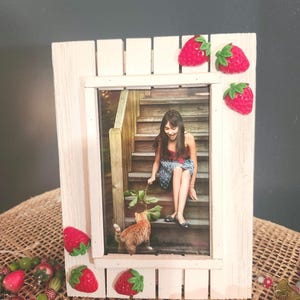 May include: White wooden picture frame with a photo of a woman sitting on stairs with a cat. The frame is decorated with red strawberry embellishments.