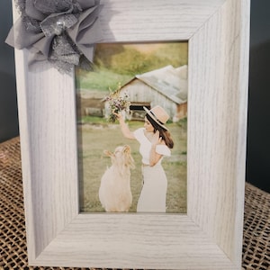 Puede incluir: Un marco de fotos blanco con una flor de tela gris en la esquina superior izquierda. El marco contiene una foto de una persona con un vestido y un sombrero blancos, sosteniendo un ramo de flores, con una cabra blanca en primer plano.