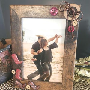 Puede incluir: Marco de fotos rústico de madera con una fotografía de una pareja bailando. El marco está decorado con adornos de botas de vaquero rojas y marrones y botones. Un lazo decorativo y botones adornan la esquina superior derecha. Acabado envejecido.