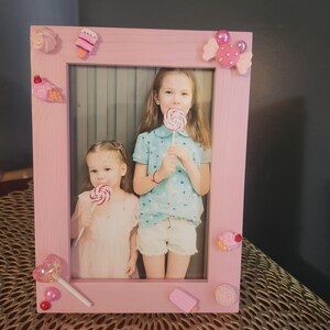 Pode incluir: Moldura de foto rosa com uma foto de duas meninas segurando pirulitos. A moldura é decorada com enfeites temáticos de doces rosa e branco.