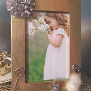 May include: Gold picture frame with a photo of a young girl in a white dress, decorated with a silver bow, a key charm, a flower charm, and a white fur pom-pom.