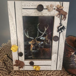 Könnte beinhalten: Ein weißer Holzbilderrahmen mit rustikalem Finish. Der Rahmen enthält ein Hirschbild und ist mit verschiedenen Tiermotiven verziert, darunter ein Bär, ein Eichhörnchen, ein Elch und ein Stinktier. Der Rahmen ist mit gelben und braunen Knöpfen verziert.