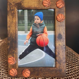 May include: Wooden picture frame with a distressed finish, adorned with small basketball accents. The frame holds a photo of a child playing basketball, wearing a blue beanie, orange vest, and black pants. The frame is displayed on a woven surface.
