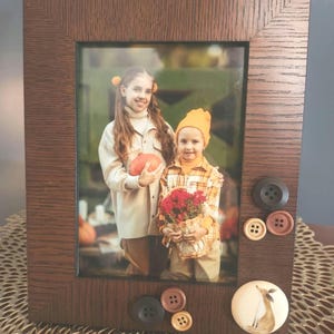 Op de afbeelding: Een bruin houten fotolijst met een foto van twee kinderen in een herfstdecor. De lijst is versierd met knopen en een keramiek vosje.