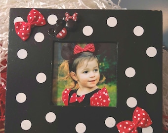 May include: Black and white polka dot picture frame with red bows and Minnie Mouse embellishments. A photo of a young girl wearing a red and white polka dot dress is in the frame.
