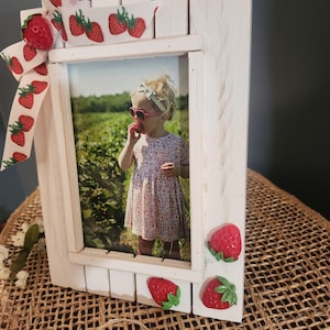 May include: A white wooden photo frame featuring a picture of a child eating a strawberry. The frame is adorned with a strawberry-printed ribbon and artificial strawberries. The frame sits on a woven surface.