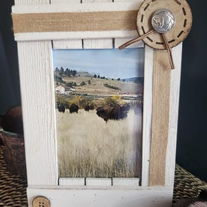 May include: White wooden picture frame with a rustic design. The frame has a brown burlap border and a brown leather button with a silver buffalo silhouette in the top right corner. The frame is holding a photo of a grassy field with a herd of buffalo in the distance.