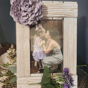 May include: A whitewashed wooden picture frame with a photo of a mother and daughter kissing. The frame is decorated with a purple flower and a small bouquet of purple flowers.