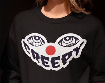 May include: A black long-sleeve crewneck sweatshirt featuring a graphic print of two stylized eyes, a solid red circle nose, and the word CREEPY in bold dark blue letters. The design is centered on the chest area of the garment.