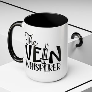 May include: White coffee mug with a black handle and black rim. The mug has black text that reads "The Vein Whisperer" with a stylized image of a syringe.