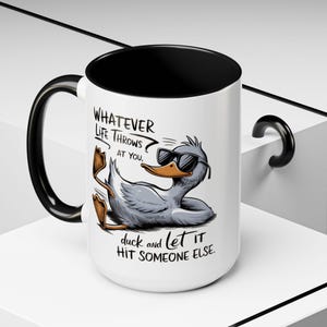 Puede incluir: Taza de cerámica blanca con asa y borde negros. La taza presenta un pato de dibujos animados con gafas de sol y el texto "WHATEVER LIFE THROWS AT YOU, duck and let IT HIT SOMEONE ELSE."