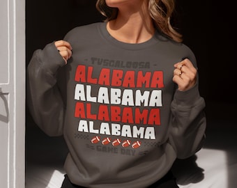 May include: A dark gray sweatshirt with the text "TUSCALOOSA" and "ALABAMA" repeated four times in red and white. The text "GAME DAY" is below the word "ALABAMA" with four footballs.