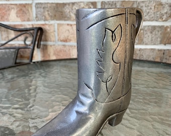 May include: A silver-coloured cowboy boot-shaped mug with a handle. The boot features etched decorative patterns and a pointed toe. The mug is made of a metallic material and sits on a glass table.
