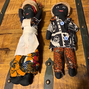 May include: Two handmade black cloth dolls. One doll wears a straw hat, a white apron, and a colorful patterned skirt. The other doll is dressed in a black and white patterned shirt, orange patterned pants, and a straw hat.