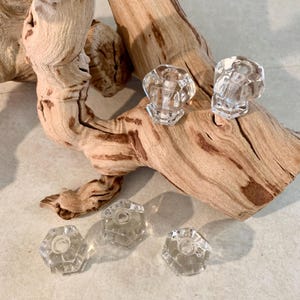 May include: Clear, faceted glass cabinet knobs with a hexagonal design. Two knobs are attached to a piece of driftwood, while three additional knobs are scattered below. The knobs are transparent, reflecting light.