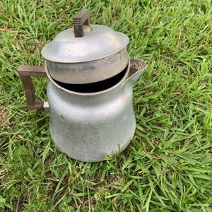May include: An aluminum coffee percolator with a lid and handle. The percolator is silver and has a dark brown handle and lid handle. The percolator is sitting on green grass.