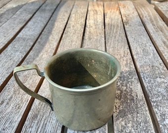 May include: An aged metal cup with a handle, showing signs of wear and a tarnished finish. The cup has a greenish tinge in places. It rests on a weathered wooden surface, with a brick wall visible in the background.