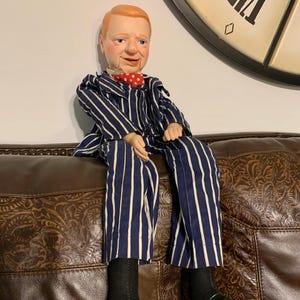 May include: A vintage ventriloquist doll with red hair, blue eyes, and a painted face. The doll wears a blue and white striped suit with a red polka dot bow tie, black socks, and black shoes. The doll is seated on a brown leather couch.