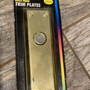 May include: A brass finish trim plate with a rectangular shape and a central button. The packaging is black with the words "BRASS FINISH TRIM PLATES" and a guarantee seal. The plate has four screw holes.