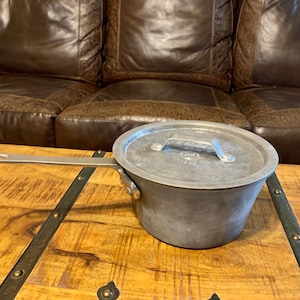 May include: A vintage metal saucepan with a lid and long handle. The pot is a dull silver color and sits on a wooden table. The handle is attached with rivets. The lid has a small handle for easy lifting. The pot is likely used for cooking.