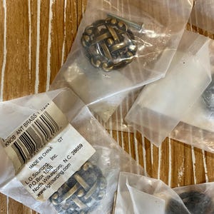 May include: Antique brass cabinet knobs with a woven design, packaged in clear plastic. Each knob includes a screw for installation. The packaging includes a label with the text "KNOB ANT BRASS".