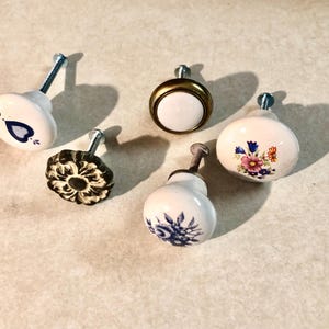 May include: A collection of five cabinet knobs featuring varied designs. One knob is white with a blue heart, one is plain white with a brass rim, one is white with a floral bouquet, one is white with blue floral patterns, and one is black with a relief.