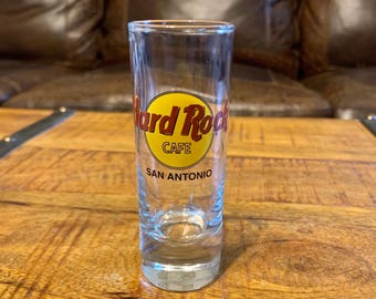 May include: A clear shot glass featuring the Hard Rock Cafe logo in yellow and red, with the text "San Antonio" below. The glass is cylindrical and rests on a wooden surface.