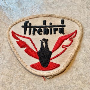 May include: A vintage Firebird patch with a cream-colored background and a red and black bird design. The word "Firebird" is embroidered in black above the bird. The patch has a stitched border.