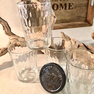 May include: Three clear glass tumblers with a textured honeycomb pattern on the exterior. A circular, dark metal lid is positioned in front of the glasses, featuring the embossed text "AMERICAN SNUFF" and "SINCE 1782" around the perimeter.