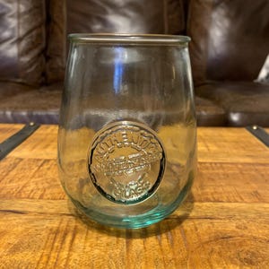 May include: A clear, stemless glass with a rounded base and a silver emblem that reads "AUTHENTIC GLASS". The glass is sitting on a wooden surface. The glass has a slight green tint.