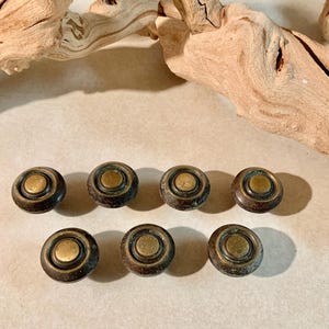 May include: Eight antique brass cabinet knobs with a circular design. Each knob features a central brass-colored circle surrounded by a darker, aged metal. The knobs are arranged in two rows of four, showcasing their vintage aesthetic.